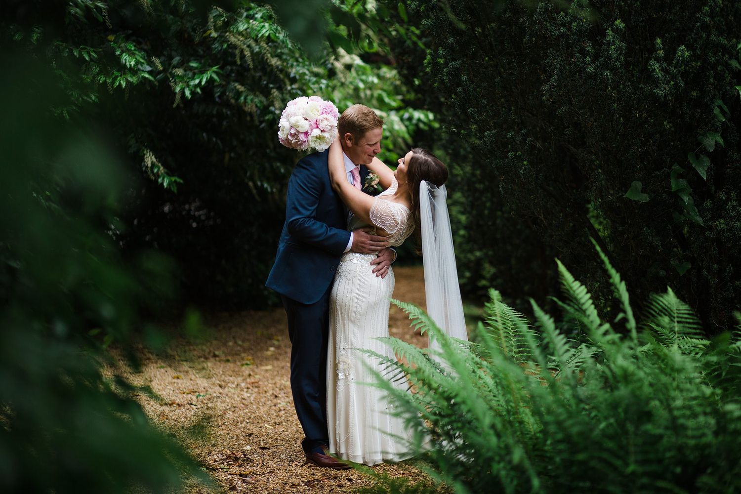 A bride and groom share a romantic embrace on a garden path surrounded by lush green ferns and trees on their wedding day.