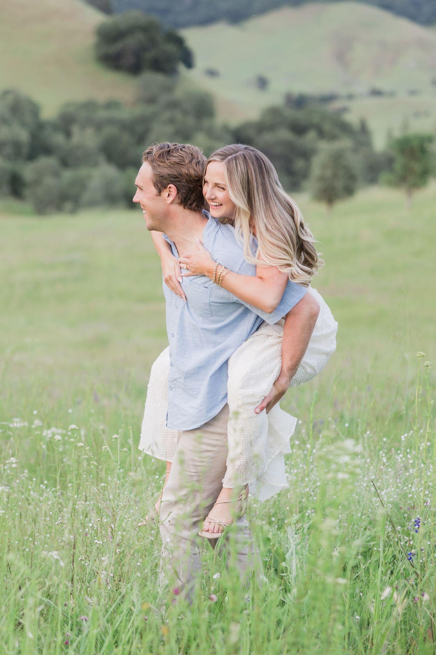 Couple sharing a kiss while showing off an engagement ring with a diamond solitaire and pearl bracelet outdoors.