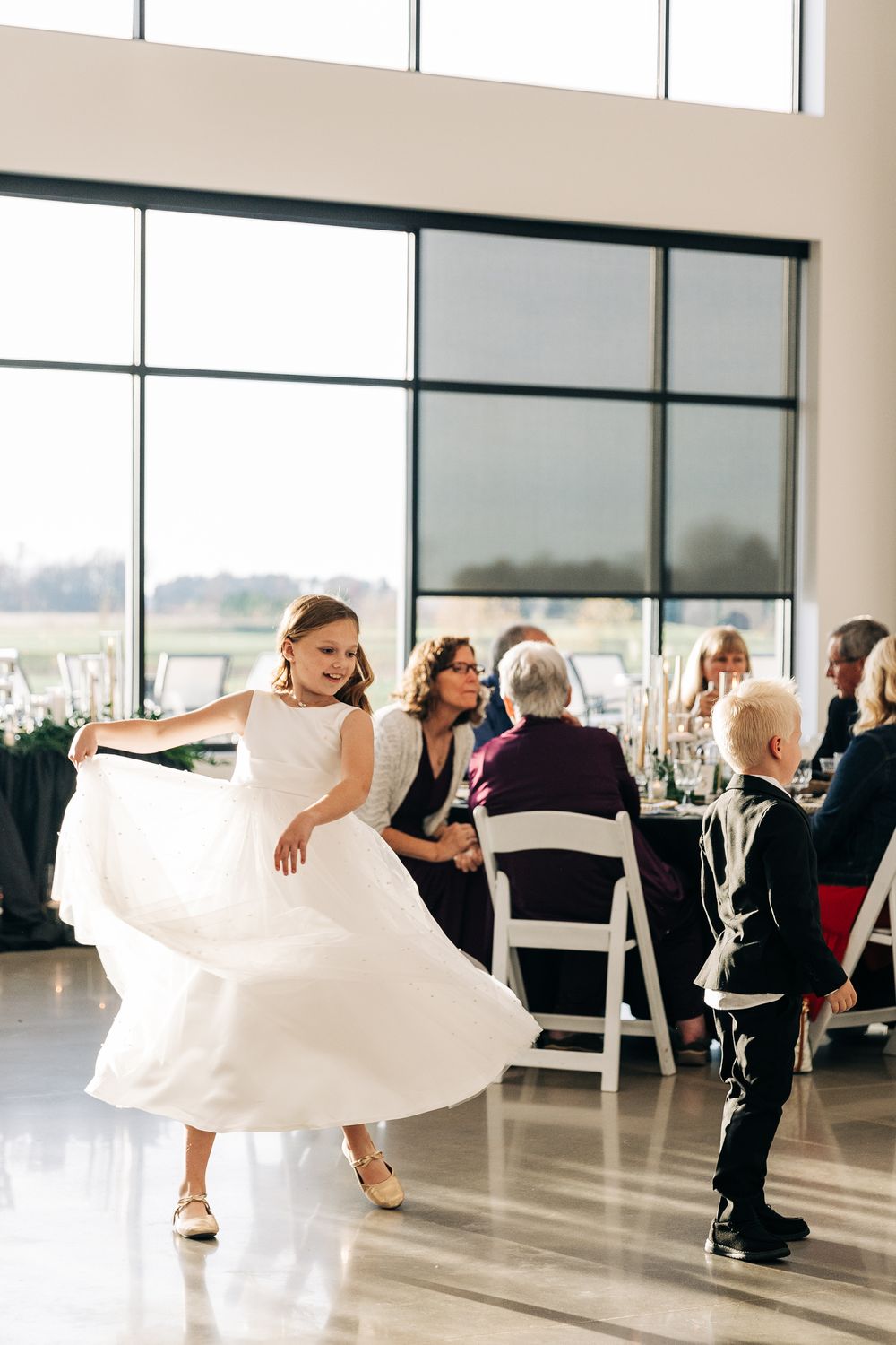 Young flower girl twirls in white dress while ring bearer watches at elegant wedding reception.