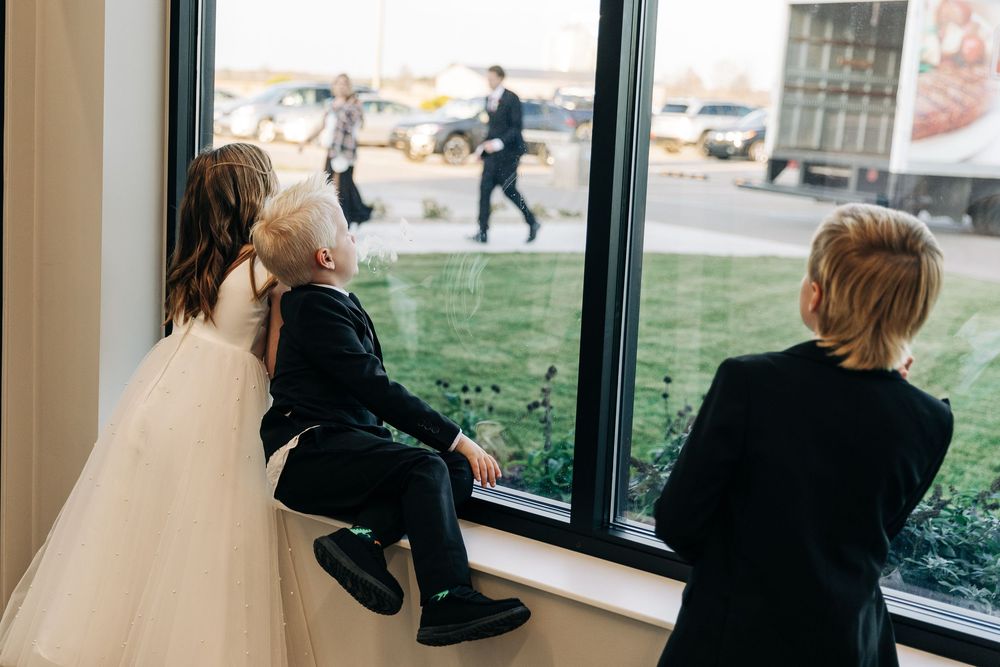 Bride in white gown with two children in formal wear looking out a large window at a wedding venue.
