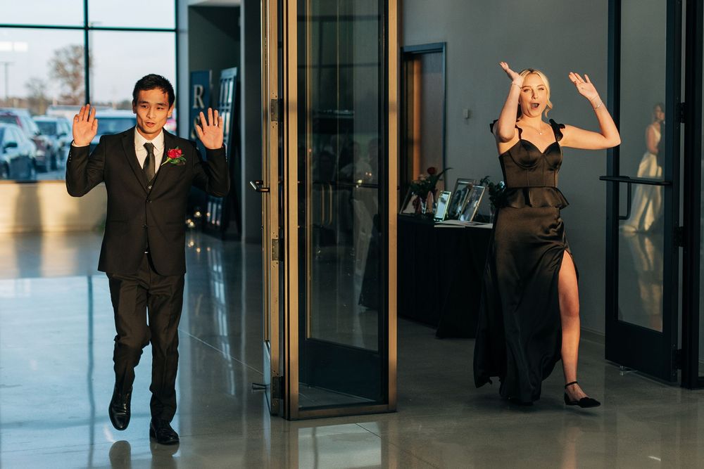 A man in a suit and woman in a black gown wave as they enter a formal event venue.