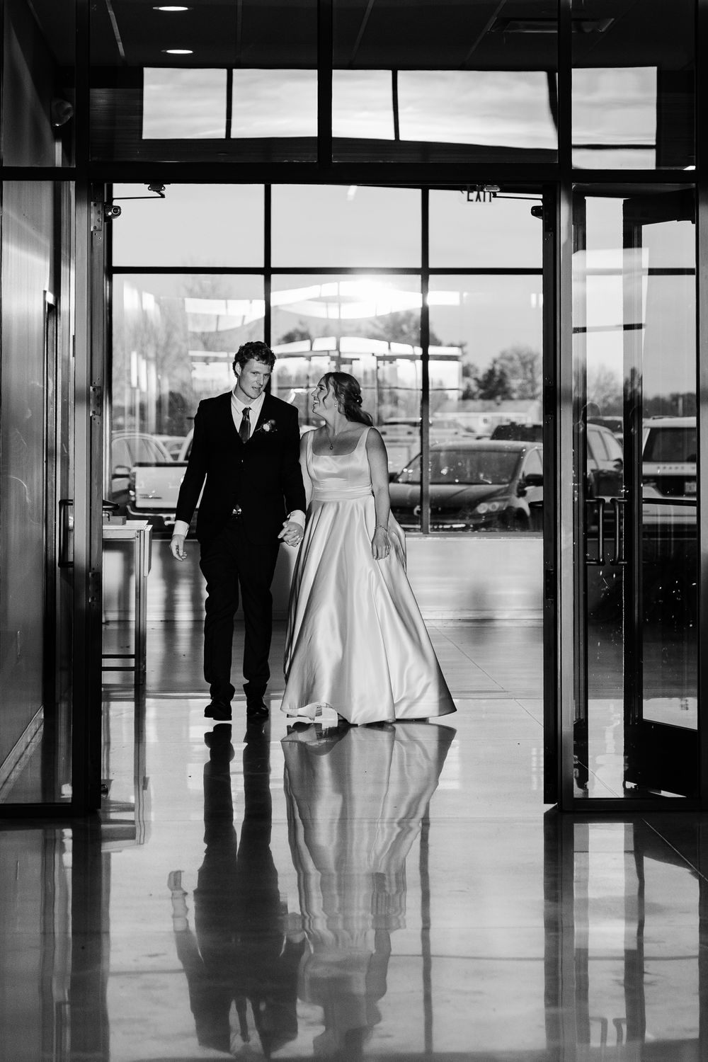 Black and white photo of bride and groom walking hand in hand through glass doors at their wedding venue.
