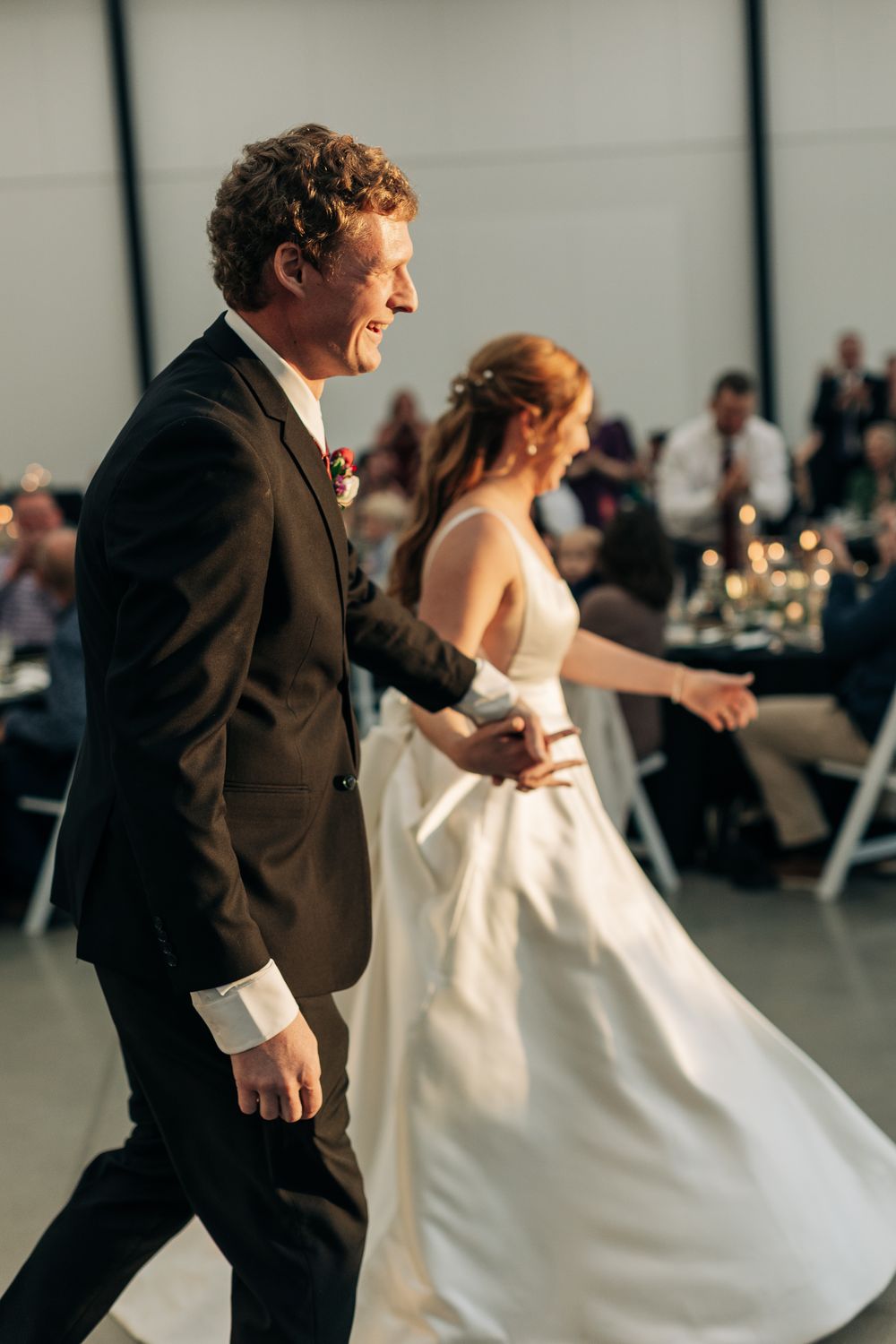 Smiling groom leads bride by hand across wedding reception dance floor in dark suit.
