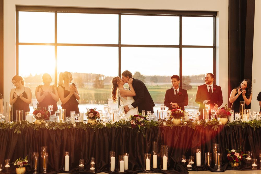 Bride and groom share a kiss at elegant wedding reception head table with candles and floral garland, golden sunset backdrop.