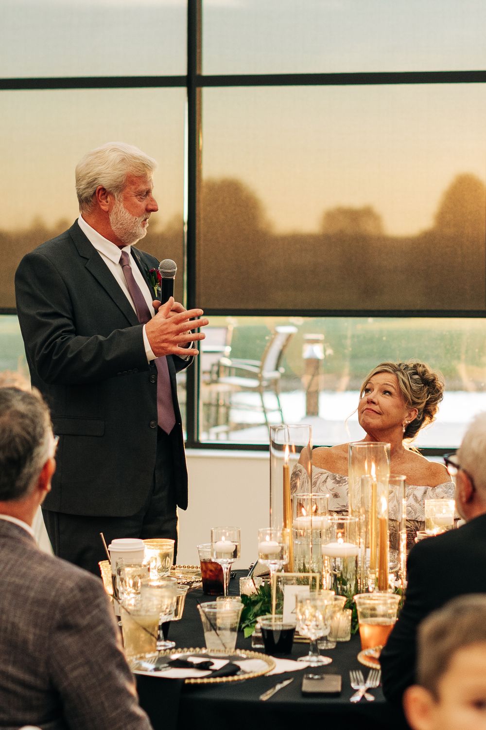 Older man in suit giving wedding speech with microphone while bride looks up at him adoringly at elegant reception.