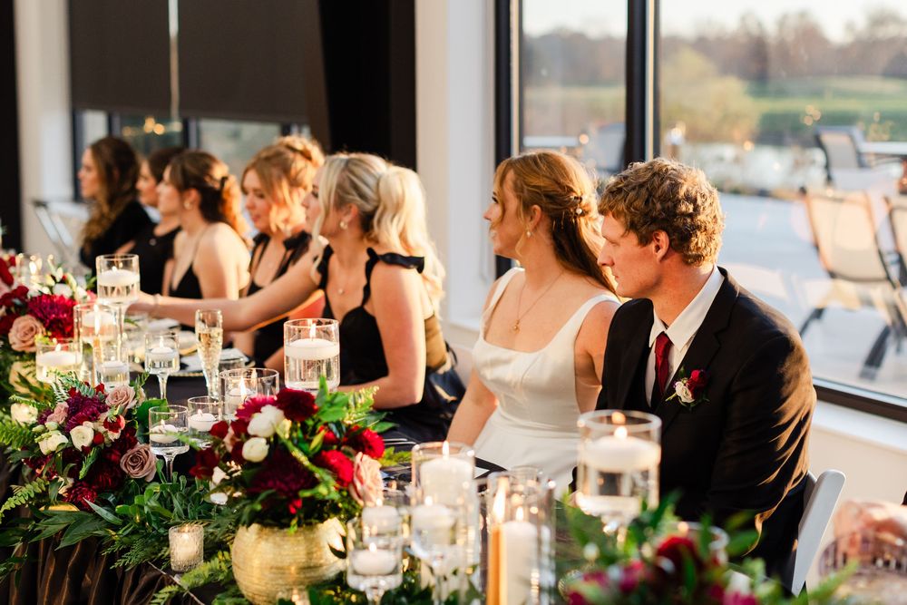 Bride and groom seated at wedding reception head table with floral arrangements and candles.