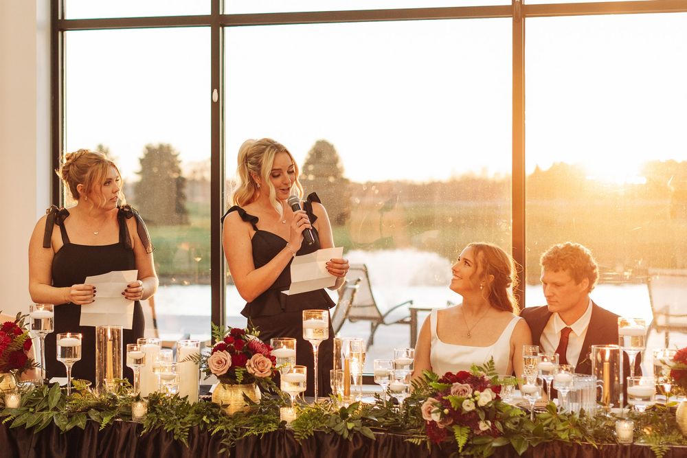 Two bridesmaids deliver heartfelt speeches at an elegant wedding reception with golden sunset light and floral table decor.
