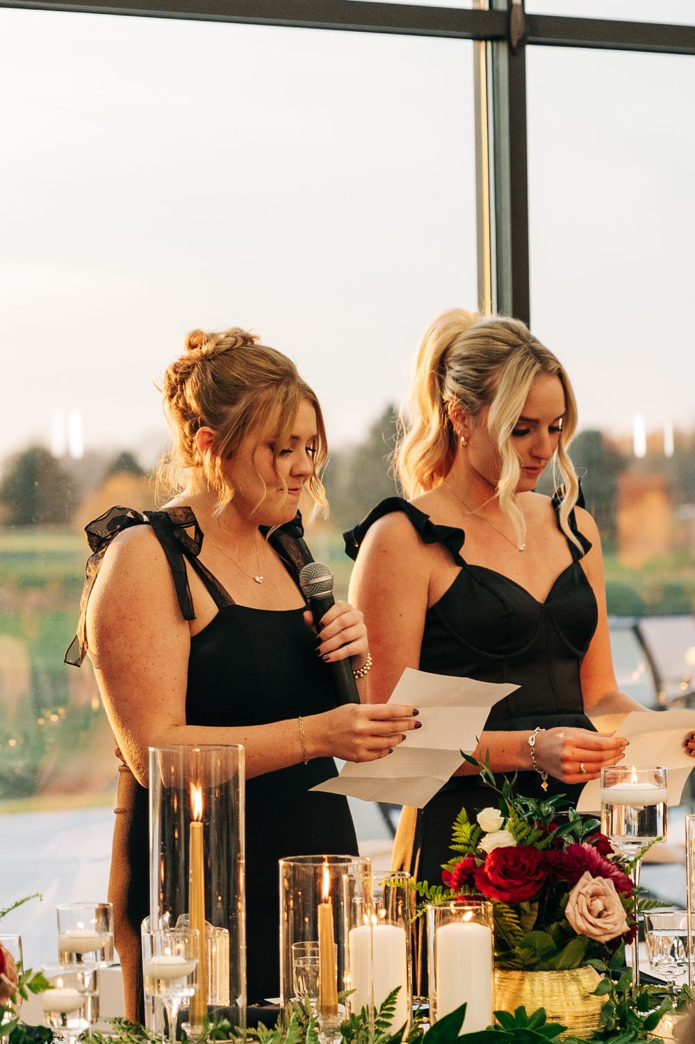 Two bridesmaids in black dresses giving a speech at a wedding reception with candles and flowers.