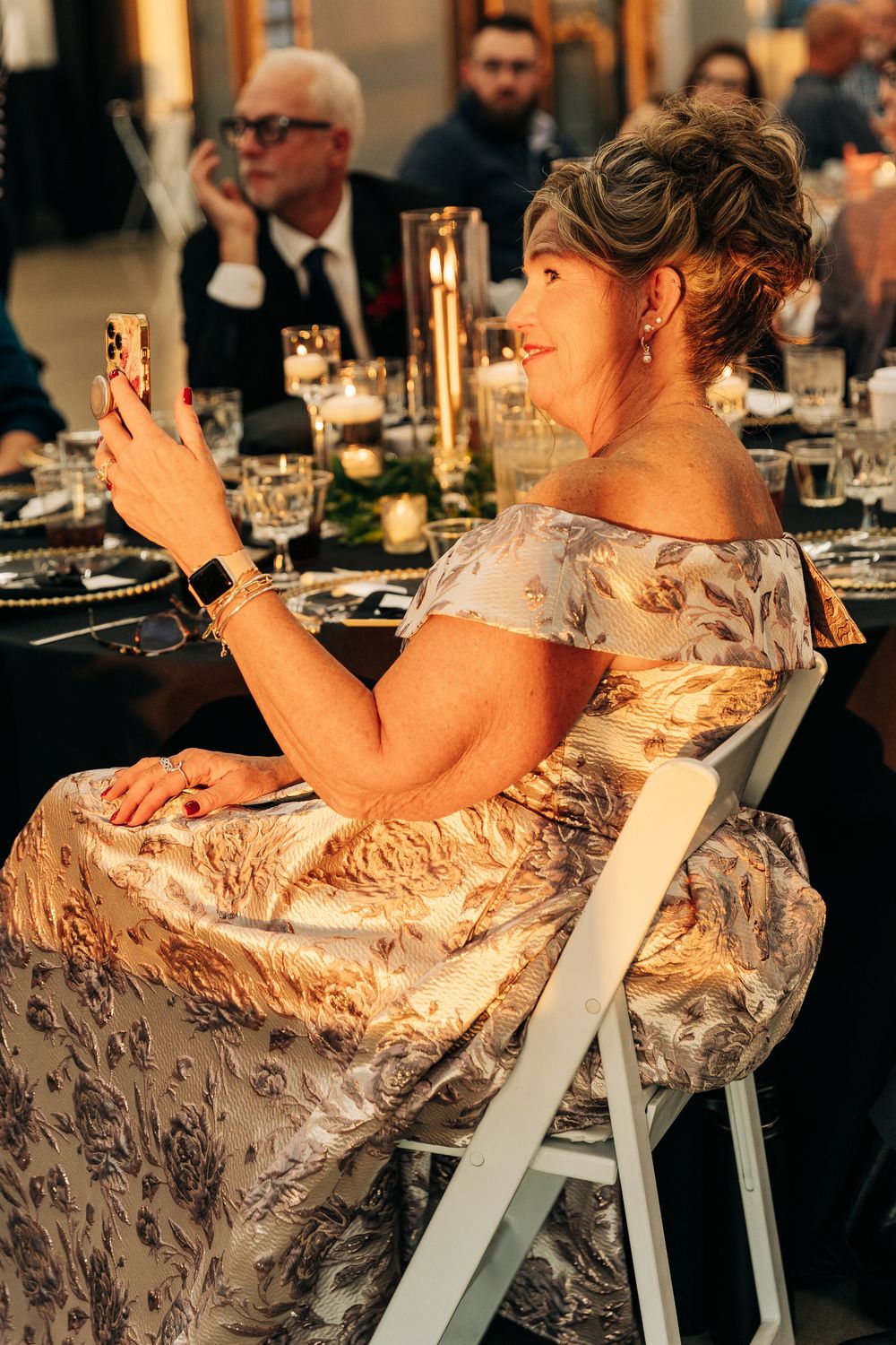 Woman in floral gown taking selfie at elegant wedding reception dinner table with candles.