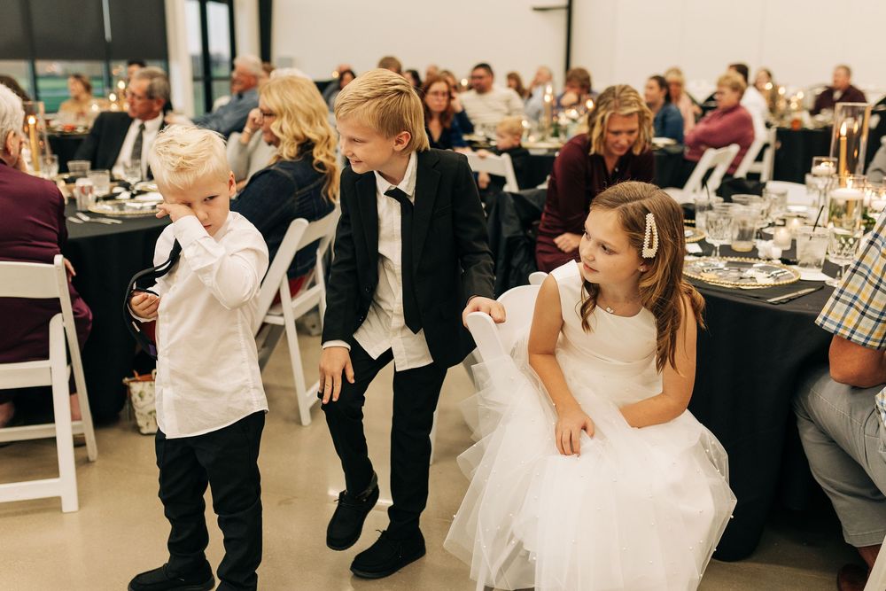 Children dressed in formal wedding attire socializing at a wedding reception with guests in background.