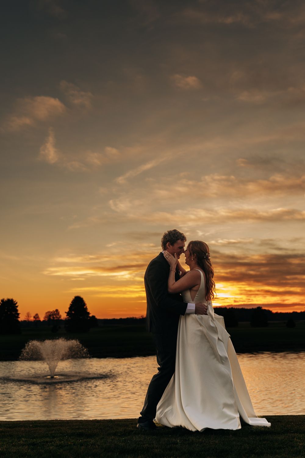 Bride and groom share a romantic moment by a pond at sunset during their wedding.
