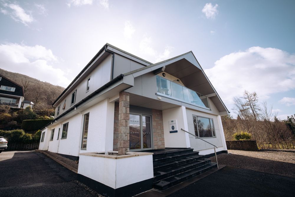 Modern white chalet-style building with glass balcony railing, stone pillars, and steps, set against a hillside backdrop.