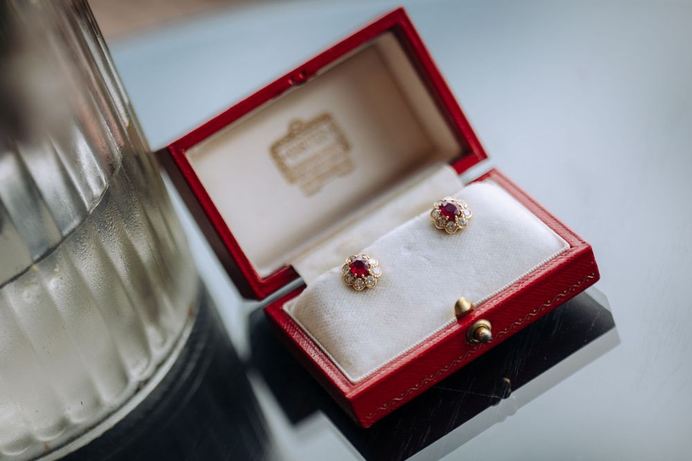 Elegant ruby and diamond stud earrings displayed in a red Cartier jewelry box on a reflective surface.