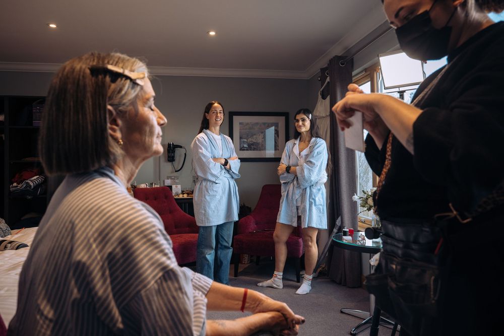 Bride getting hair done by stylist while bridesmaids in white robes watch in a cozy room.