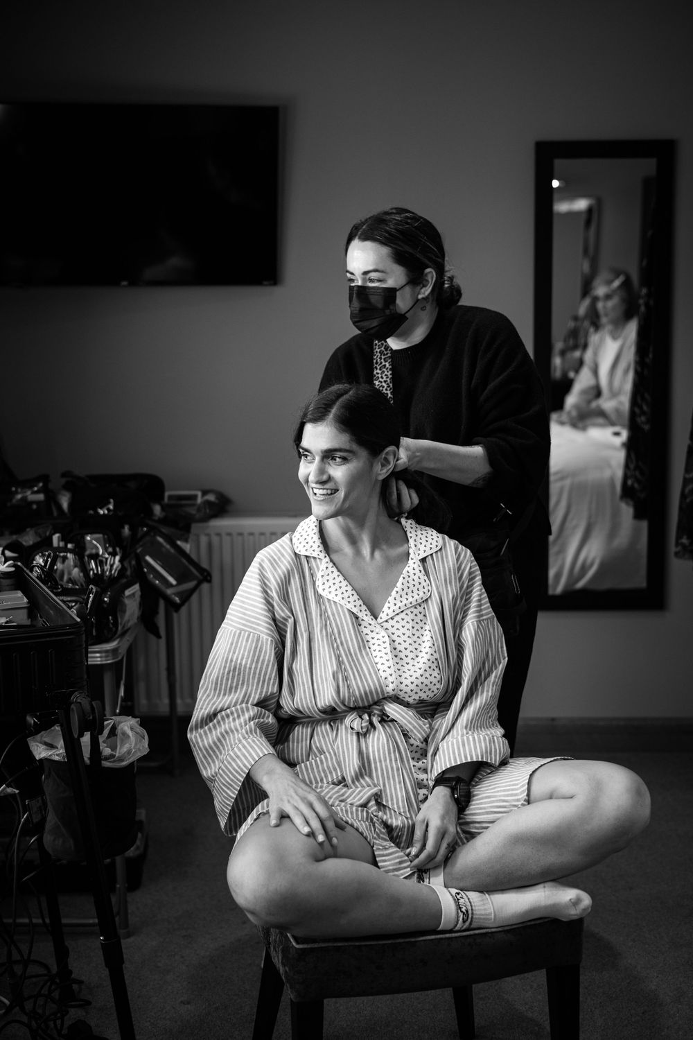 Black and white photo of a bride getting her hair done by a masked stylist while sitting cross-legged on a stool.