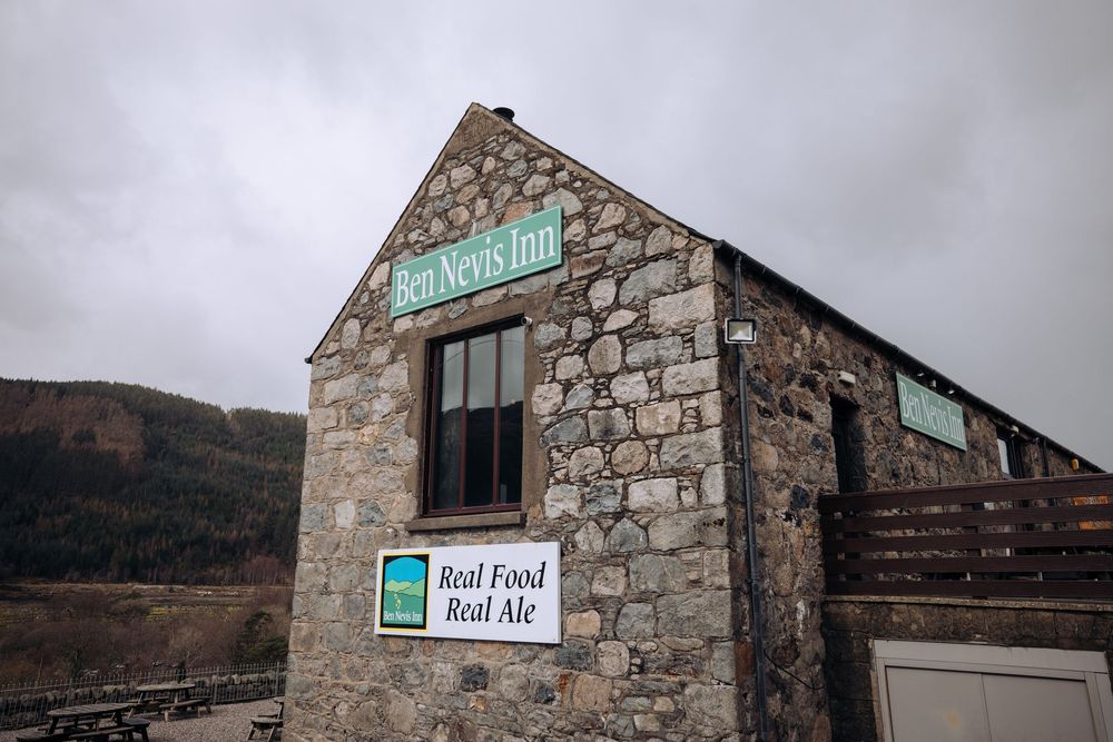 Ben Nevis Inn stone building with Real Food Real Ale sign, set against Scottish Highland hills under overcast sky.