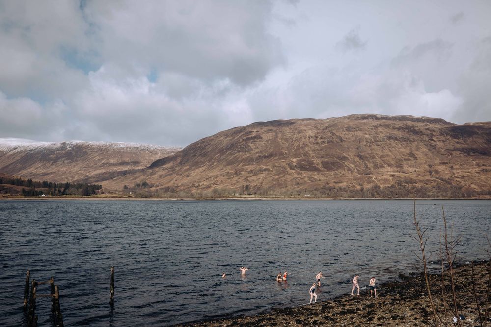 People wading in a cold Scottish loch with snow-dusted hills under cloudy skies.
