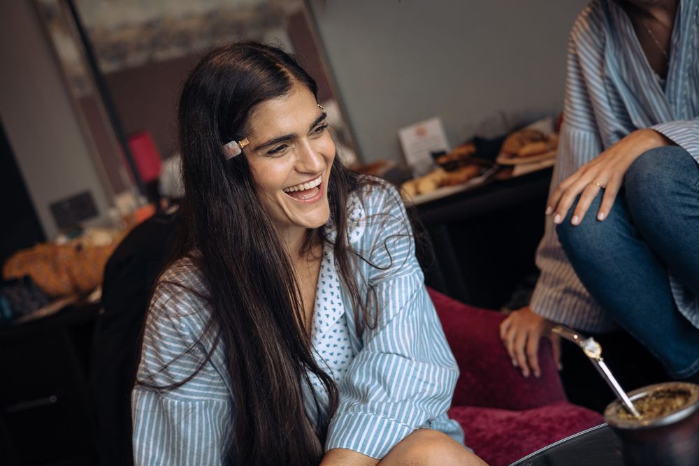 Young woman laughing joyfully while sitting in a room, wearing a blue striped shirt with a pencil in her hair.