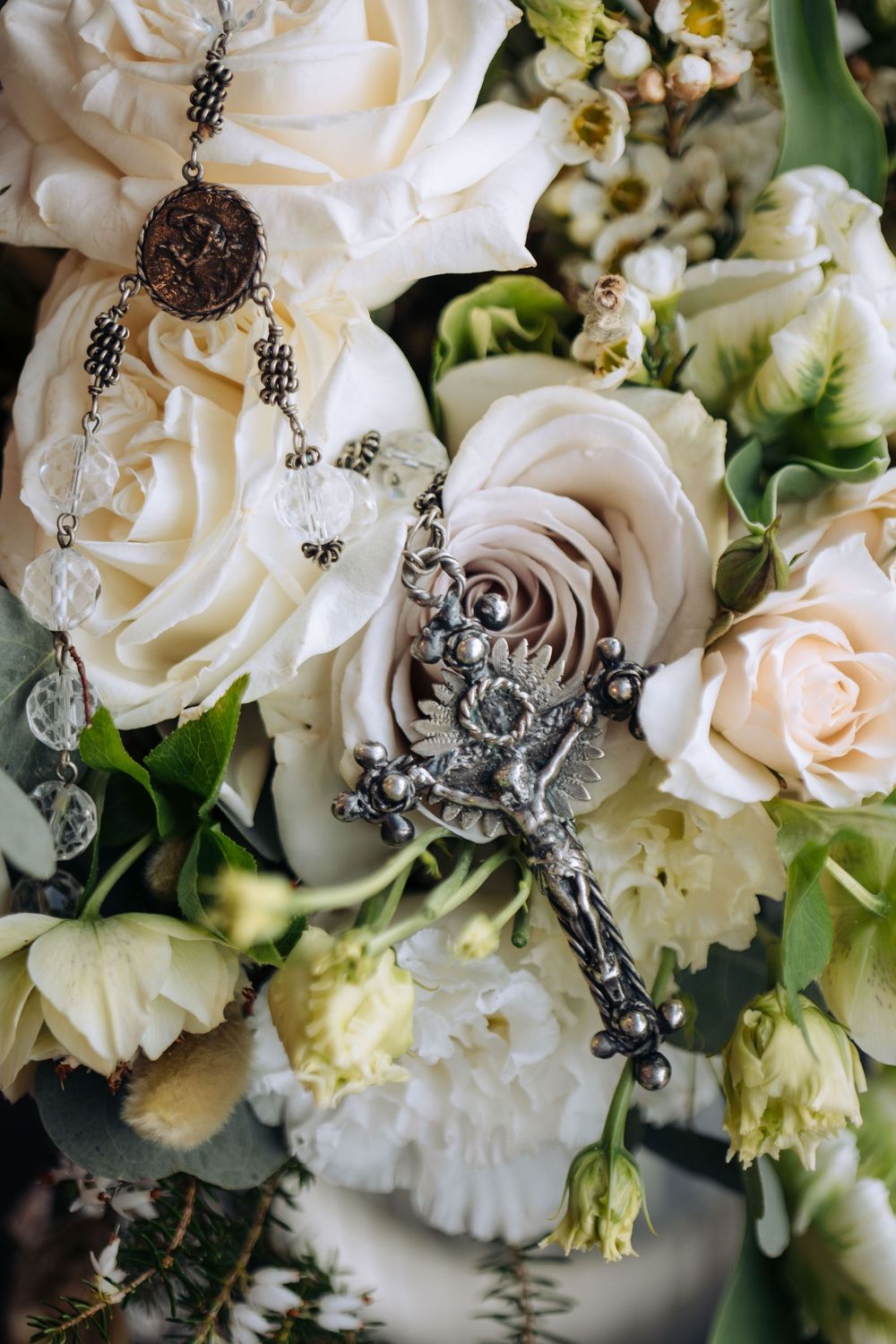 Silver crucifix rosary resting on elegant white and blush rose wedding bouquet with greenery.