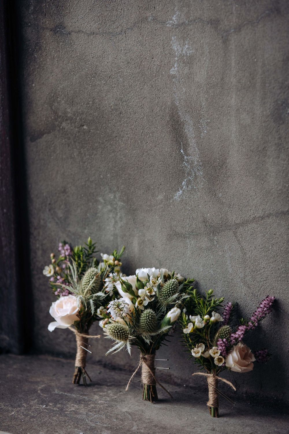 Three rustic wildflower bouquets with thistles and roses leaning against a gray concrete wall.