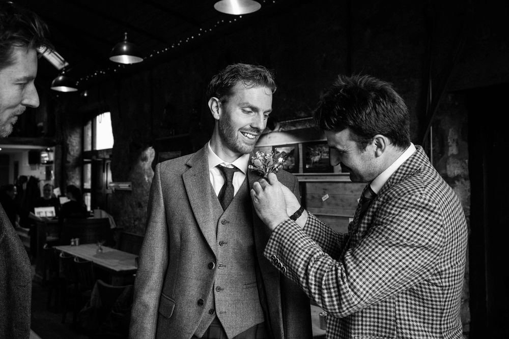Groom in tweed suit getting boutonniere pinned by groomsman in black and white wedding photo.