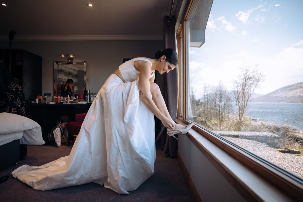 Bride in white wedding gown adjusting her shoe by a large window with scenic loch views in a hotel room.