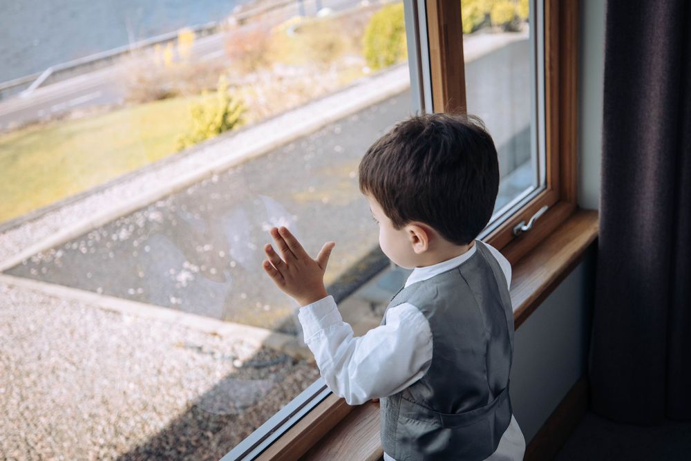 Young boy in a vest looking out a window at a scenic outdoor view, hands pressed against the glass.