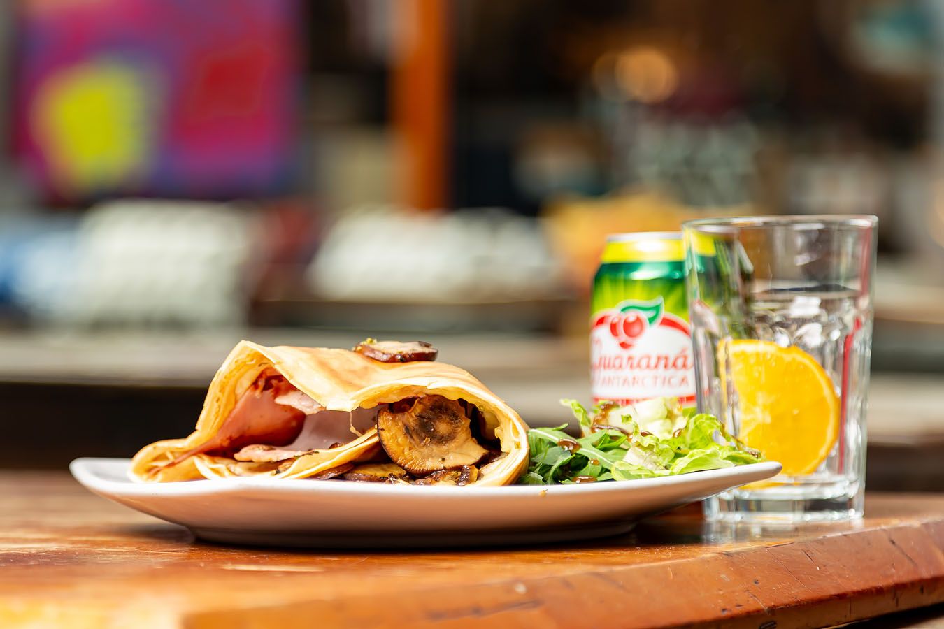 Savoury crepe with mushrooms and ham, served with a side salad, Guaraná Antarctica, and a refreshing drink on a wooden restaurant table.