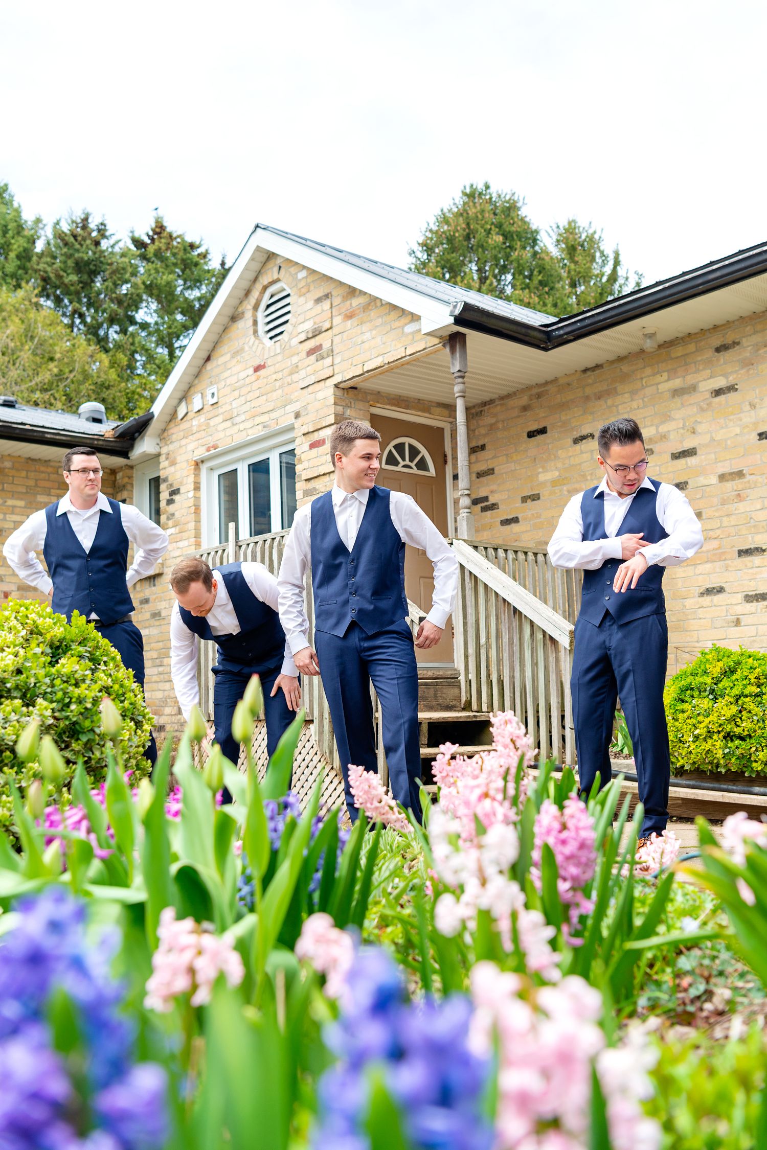 Groomsmen in navy vests and white shirts stand near colorful spring flowers outside a brick house.