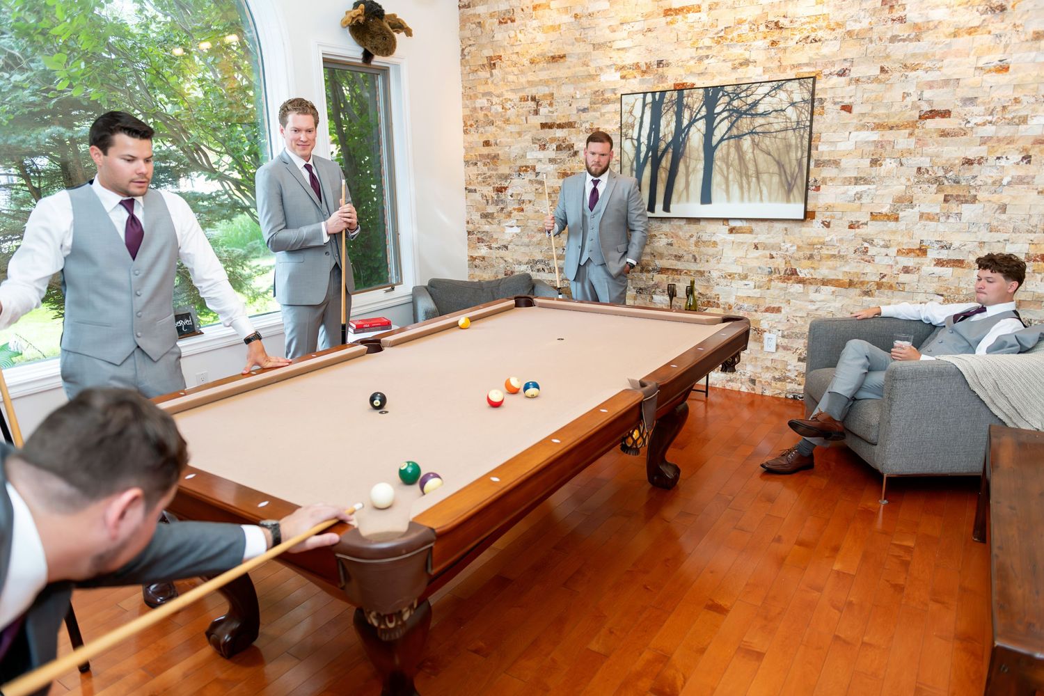 Groomsmen in gray suits playing pool in an elegant room with stone walls and hardwood floors.