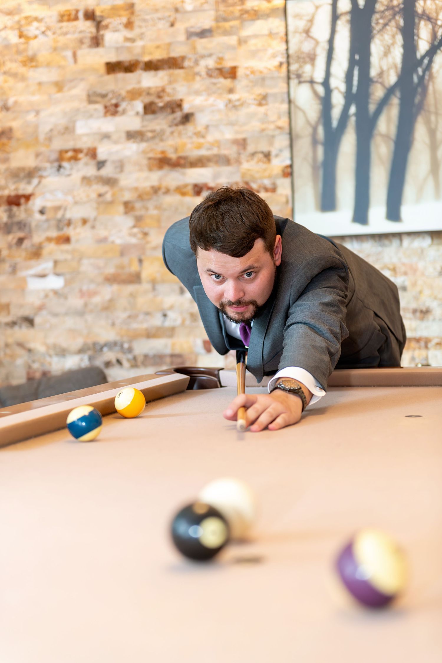 Man in suit leaning over pool table aiming cue stick at billiard balls in stylish brick-walled game room.