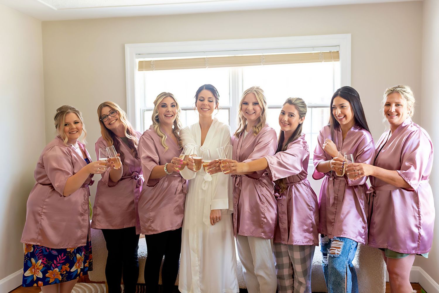 Bride in white robe surrounded by bridesmaids in pink robes holding hands together near a bright window.