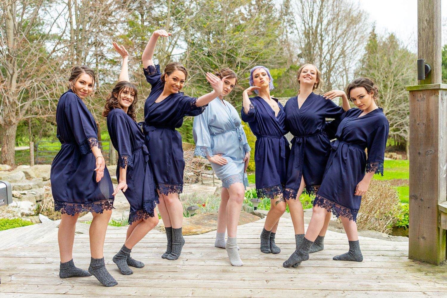 Seven bridesmaids in navy satin robes pose playfully on a wooden deck surrounded by spring greenery.