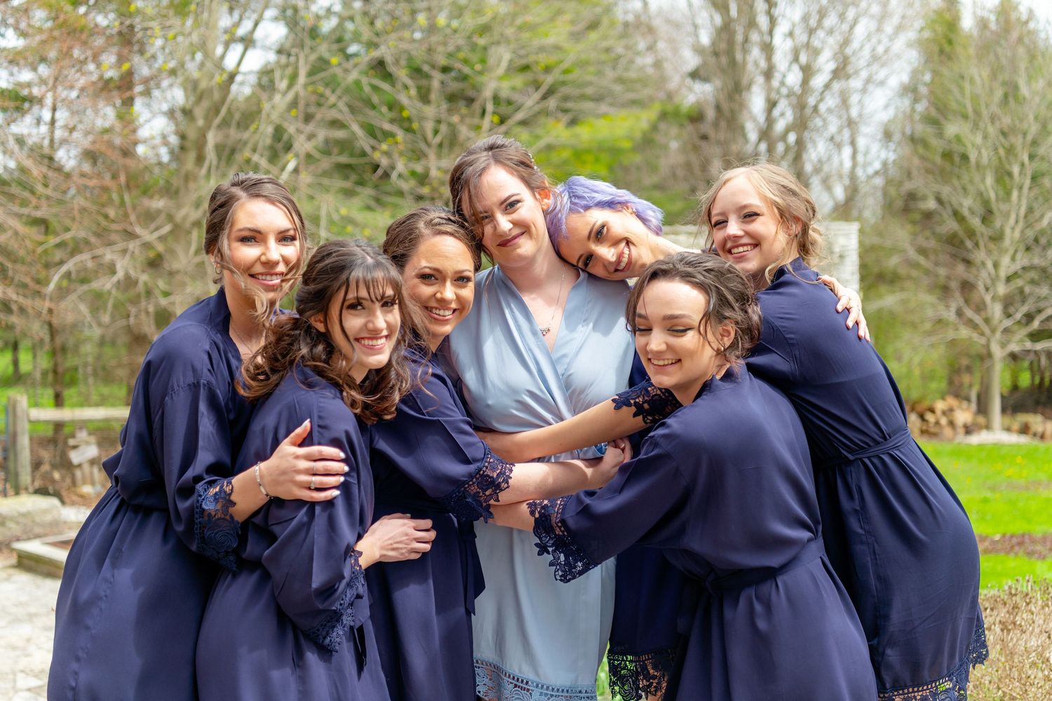 Bride in blue robe surrounded by smiling bridesmaids in navy robes hugging outdoors at a wedding venue.