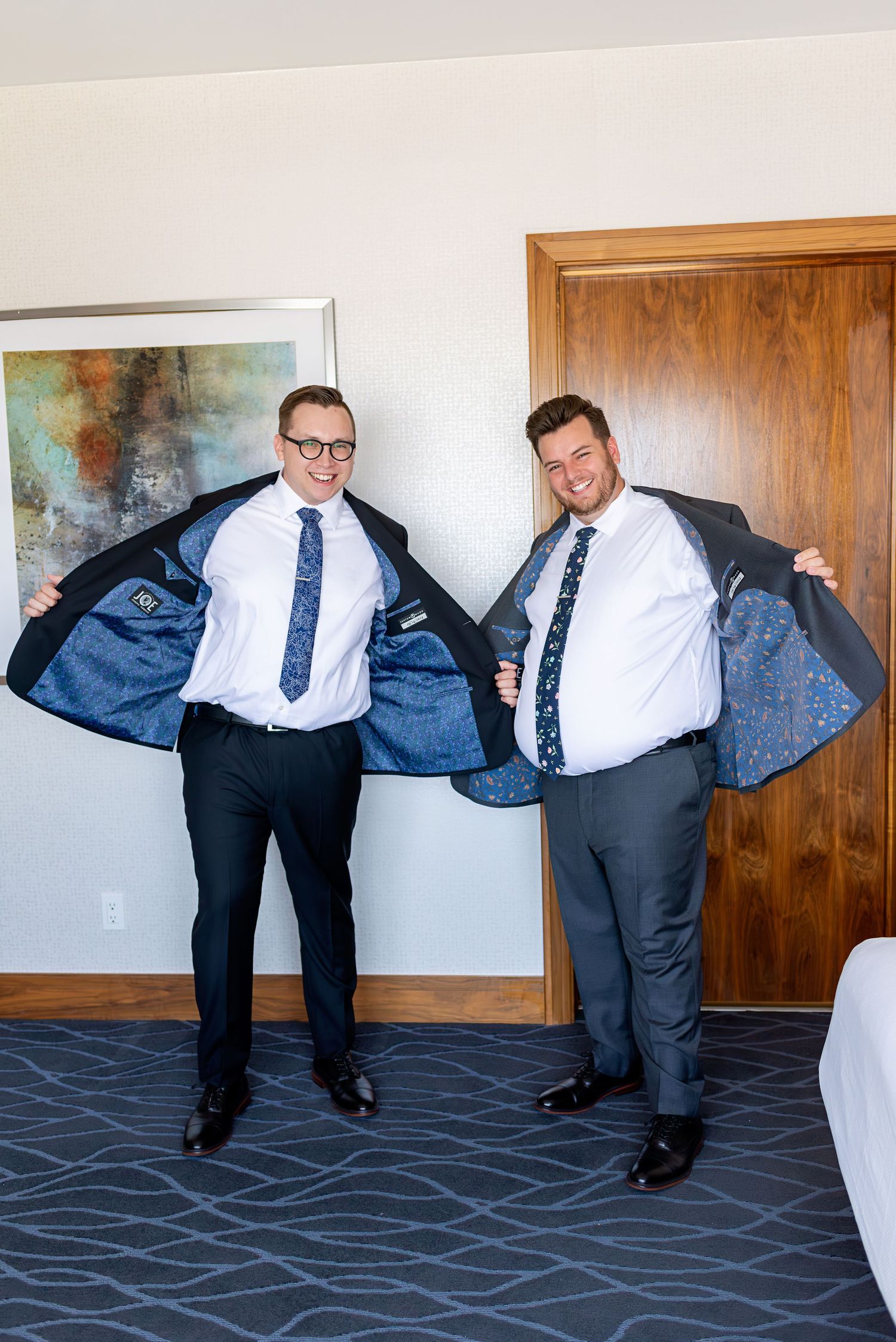 Two groomsmen in dark suits proudly showing off their jacket linings in a hotel room before a wedding.