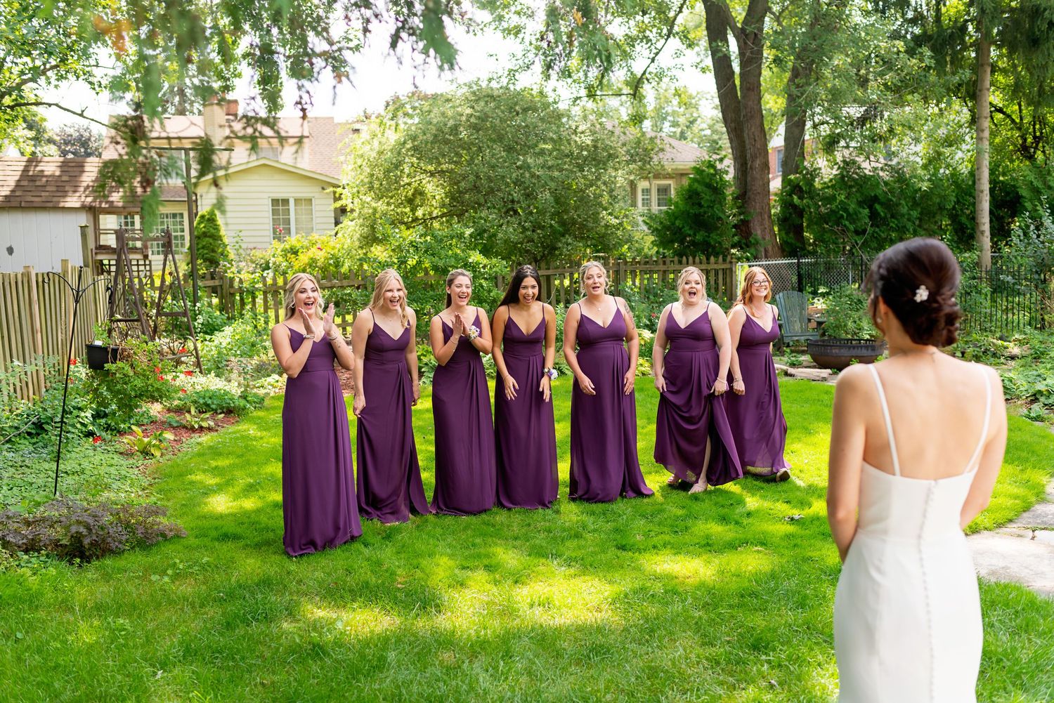 Bride in white gown reveals herself to bridesmaids in purple dresses during outdoor first look ceremony.