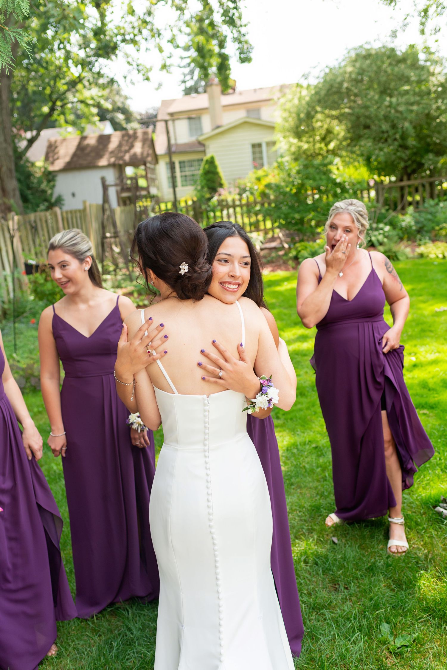 Bride in white gown hugging smiling bridesmaid in purple dress outdoors, with emotional wedding party in background.