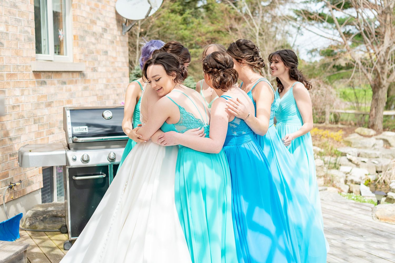 Bride in white gown shares a group hug with three bridesmaids in blue dresses on an outdoor patio.