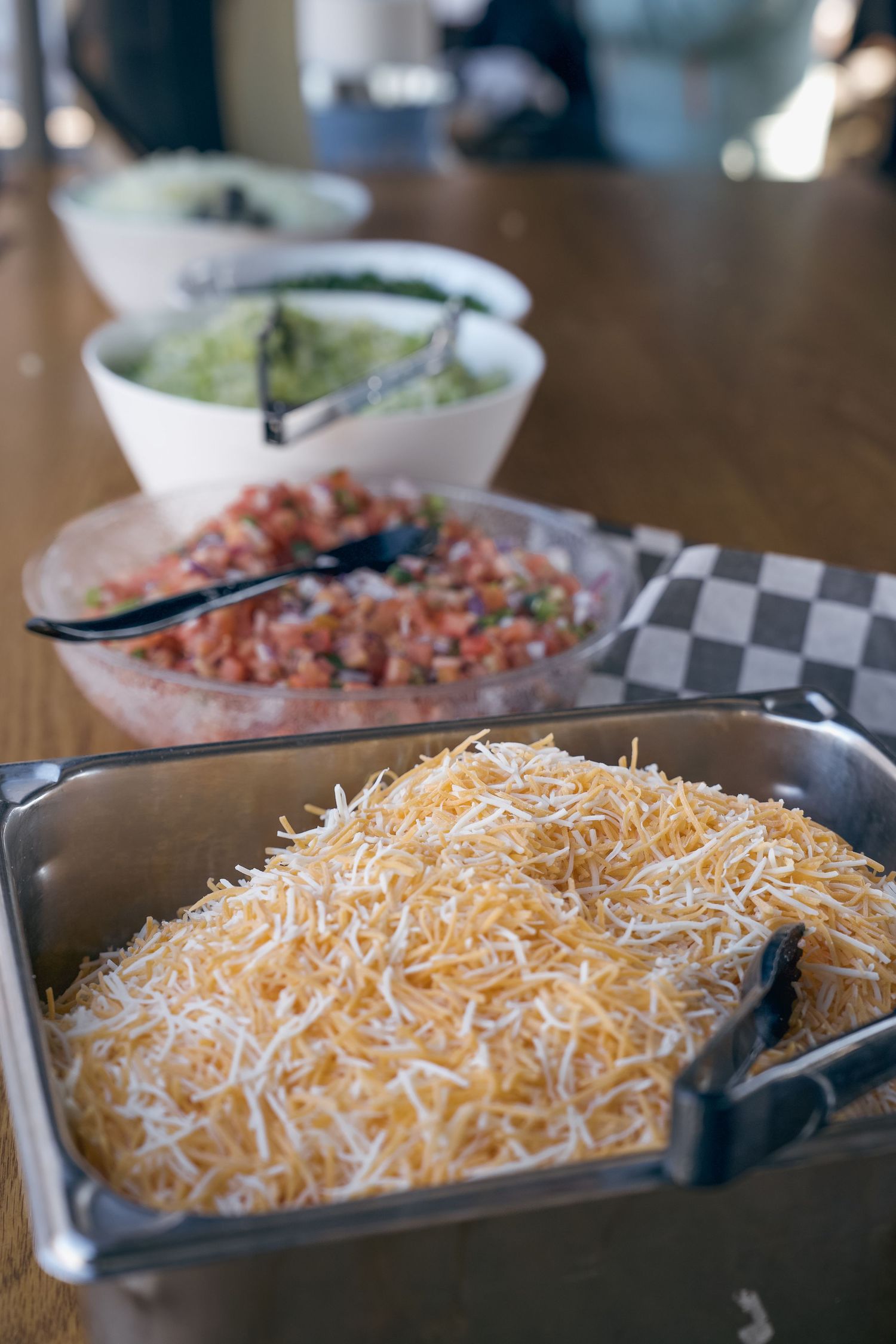 Shredded cheese in a metal tray with salsa and guacamole bowls on a wooden table at a taco bar.