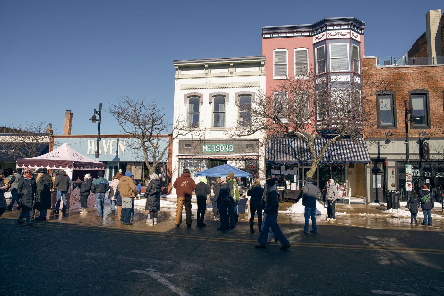 People gather outside historic brick storefronts on a sunny winter day in a small town square.