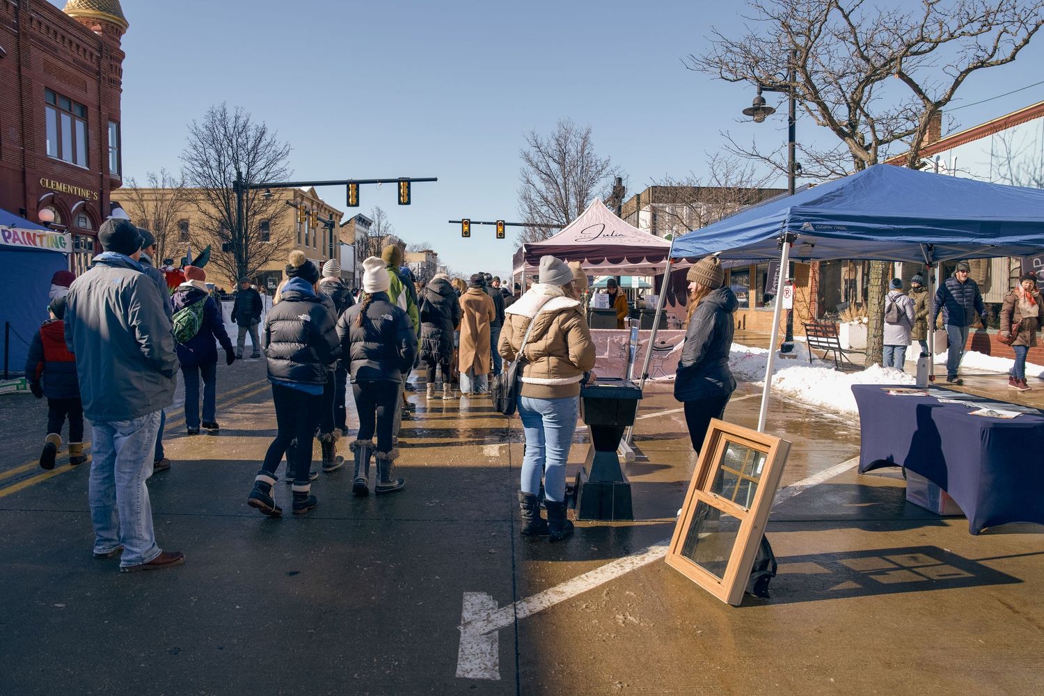 People browsing outdoor winter street market with tents and vendors on a sunny day in a small town.