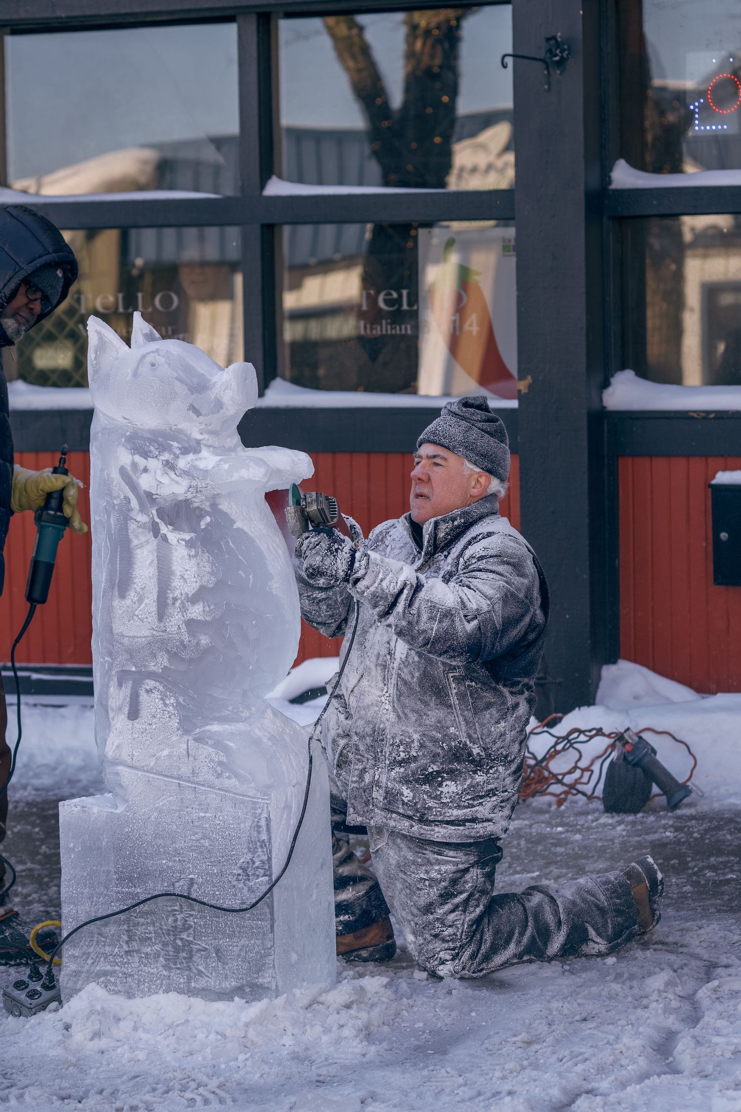 Man kneeling in snow carefully sculpting a detailed ice figure outside a restaurant in winter.