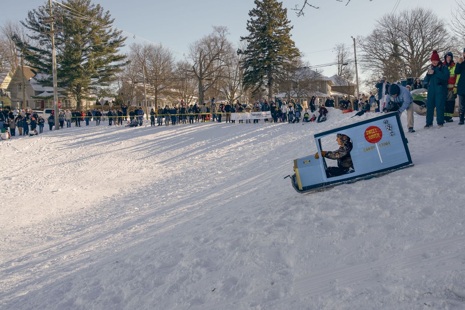 A cardboard sled races down a snowy hill during a winter festival, with spectators lining the course.