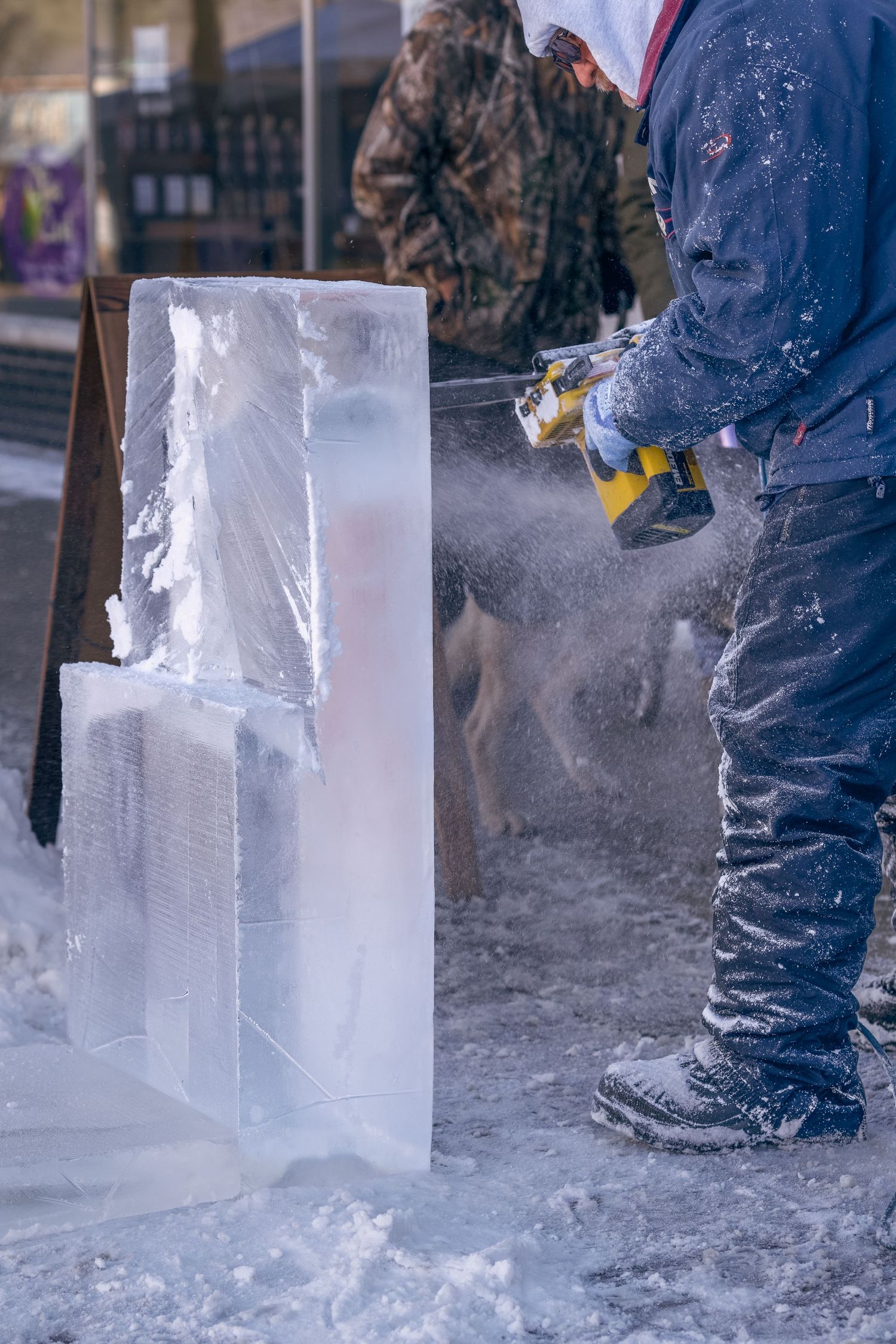 Ice sculptor uses power tool to carve large clear ice block outdoors in winter.