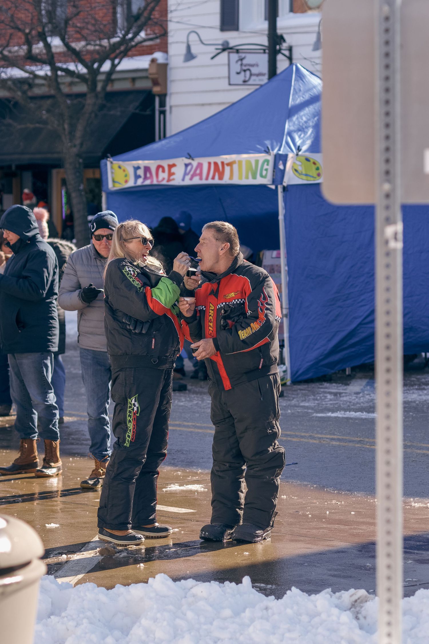 People gathered at a winter outdoor festival near a face painting tent with snow on the ground.
