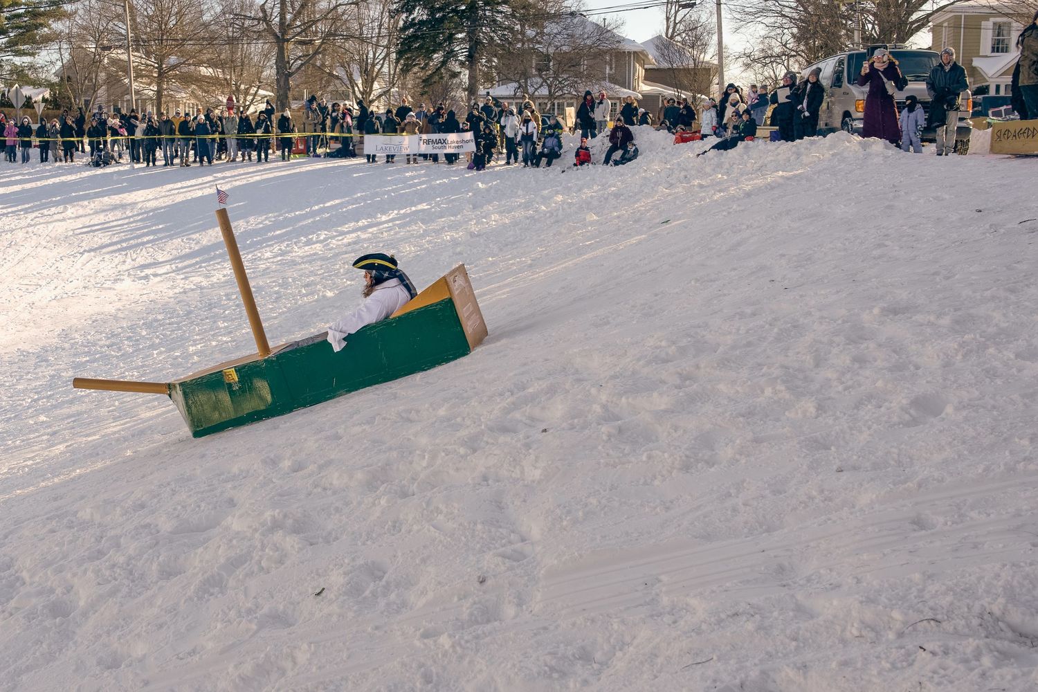 Person sledding down a snowy hill on a wooden sled at a winter festival with spectators watching.
