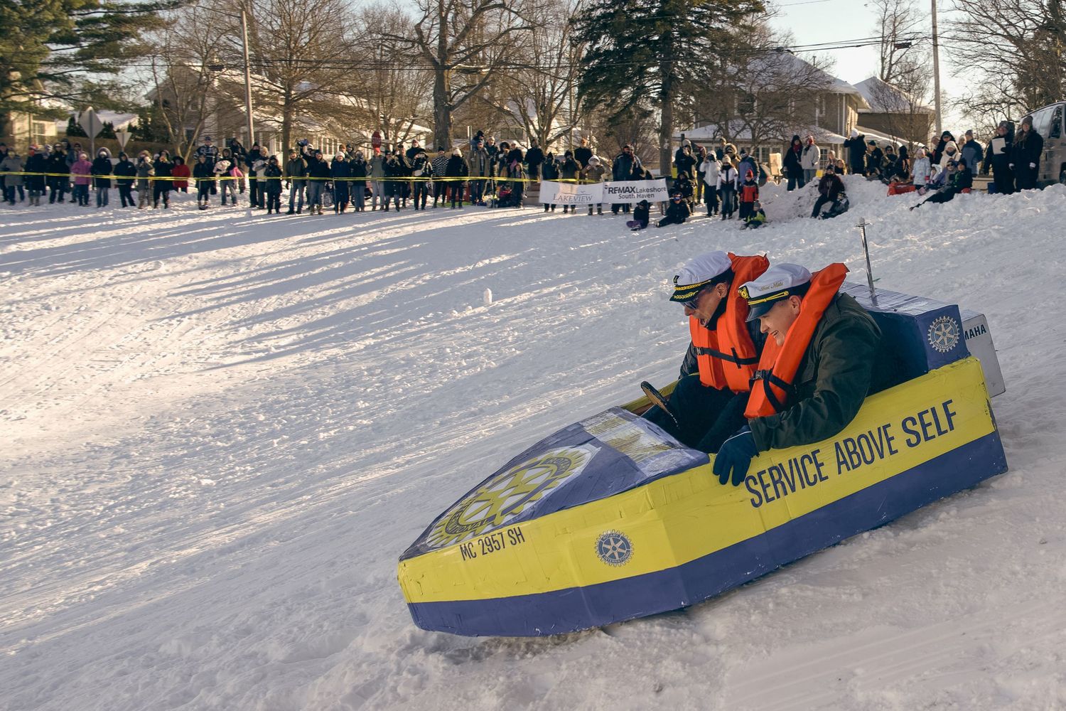 A blue and yellow sled reading 'Service Above Self' races down a snowy hill during a winter sledding competition.