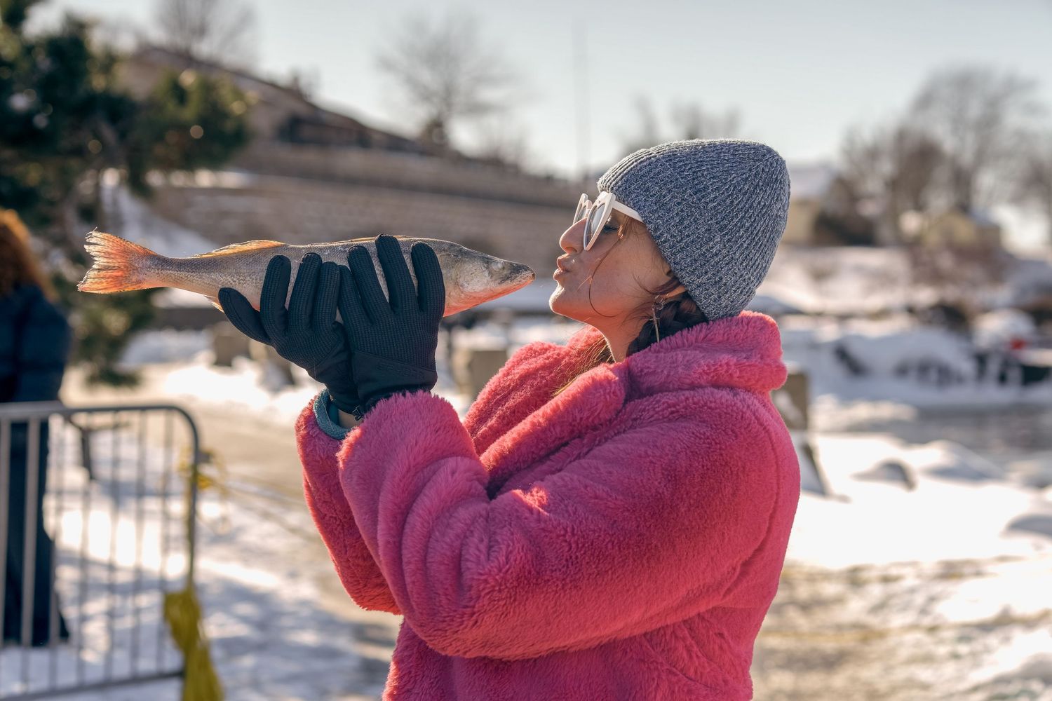 Woman in pink coat and gray beanie holds a fish outdoors in a snowy winter setting.