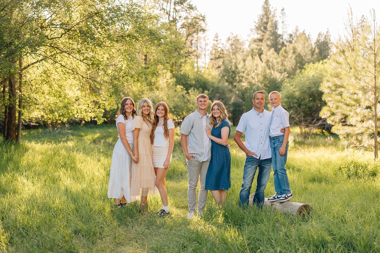 Family of six posing outdoors in a sunny green meadow surrounded by trees during a warm summer day.