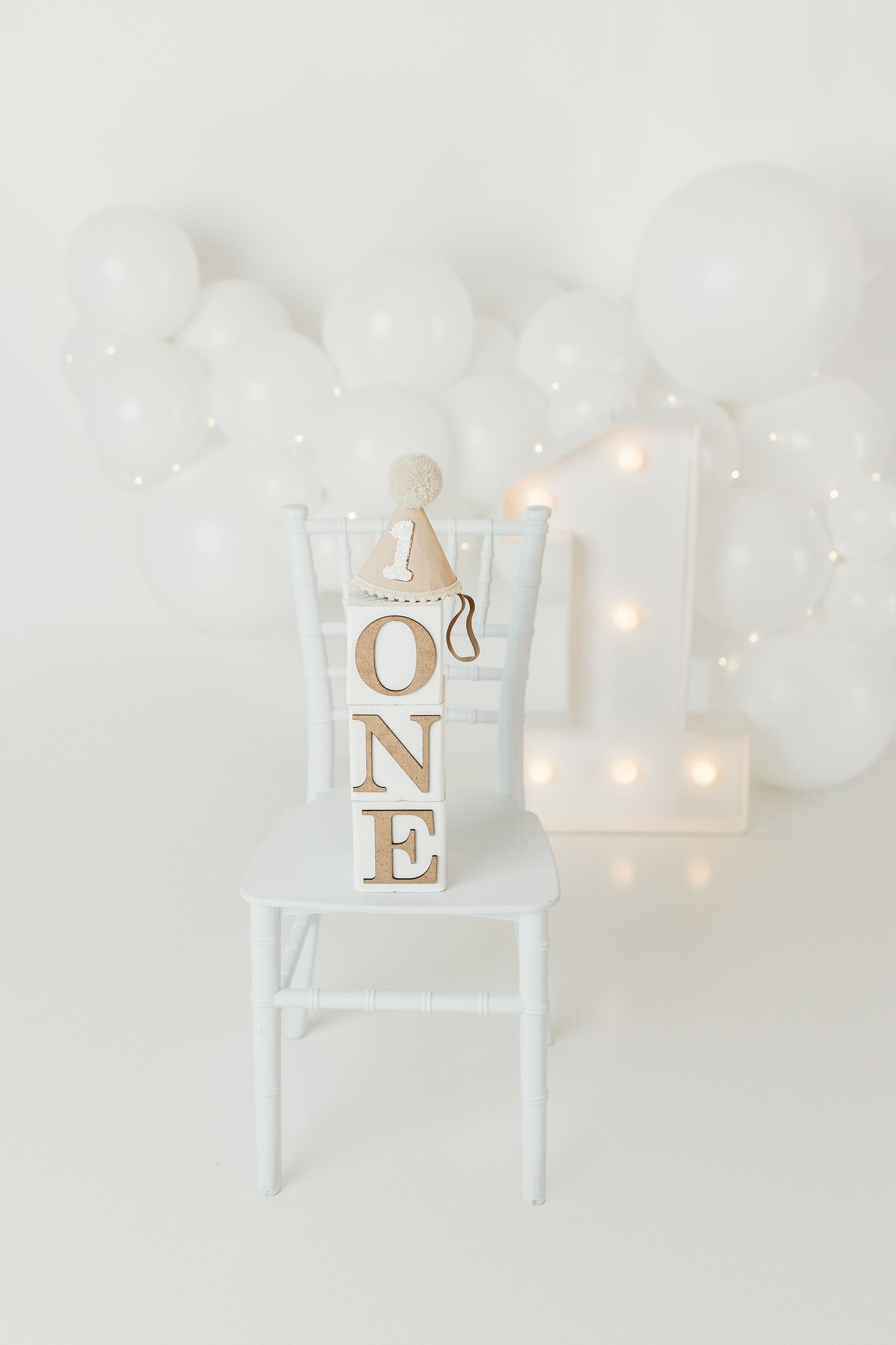 White first birthday setup with ONE blocks, party hat, lit number 1, and white balloon garland on white chair.